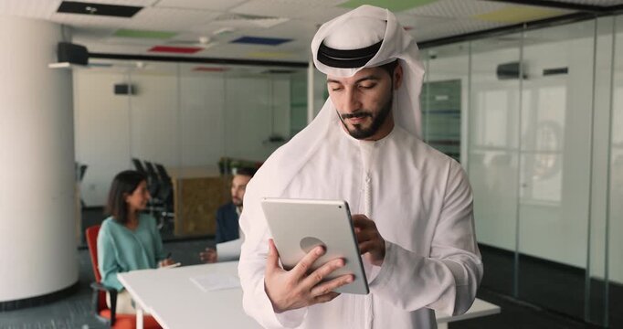 Focused young Arabian professional man typing on digital tablet pc, standing in meeting room with diverse colleagues in background, using gadget for online work business communication