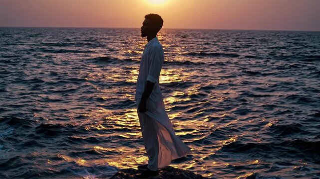 Man in a white robe standing on rocks by the ocean at sunset. Traditional clothing blowing in the wind at the seashore. Travel and contemplation concept