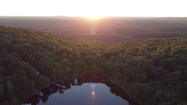 Flying over Buckskin Lake away from the sun with lens flare on a summer afternoon before sunset. Aerial of lakeside cottages and of crown land wilderness near Tory Hill in Halliburton Highlands East.