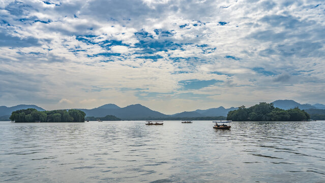 Tourist boats sail on the calm and beautiful lake. Ripples, reflections on the water. Green islands. Mountains on the horizon. Blue sky, clouds. China. Hangzhou. West Lake Xi Hu