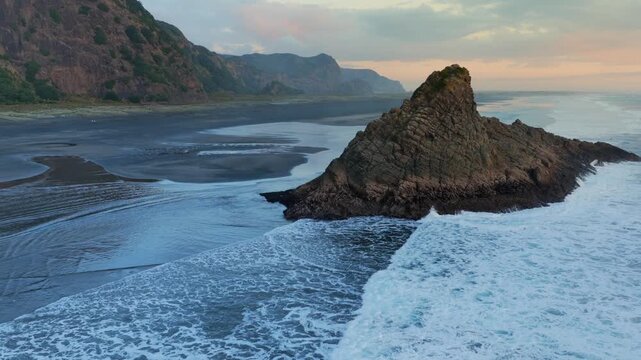 Aerial view of the Tasman sea crashing against the rocky outcrop at sunset, in Karekare Beach, Auckland, New Zealand. The waves are crashing against the rocks, eroding them.