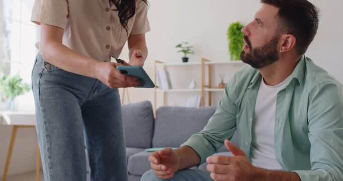 Couple arguing over smartphone messages of husband in living room. Jealous wife in anger showing phone screen with cheating SMS or chat, discussion about trust of annoyed girl and man at home