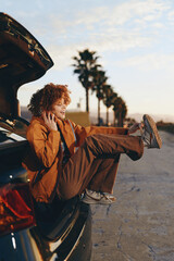 Fototapeta premium Smiling woman with curly hair sitting in car trunk wearing a brown jacket and rainbow sweater, enjoying a relaxed lifestyle outdoors with palm trees and clear sky in background.