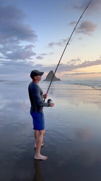 A man is surfcasting at sunset on Karekare Beach, New Zealand. He is holding a fishing rod and reel, hoping to catch fish for dinner. The sky is colorful and reflective.