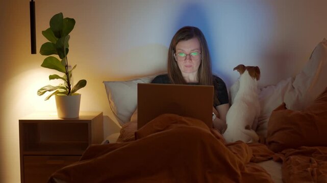 Woman wearing glasses typing on laptop in bed under blanket with small dog. Person using laptop in bedroom before sleeping. Concept of remote work online study and domestic life