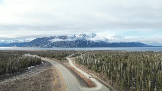 Cinematic drone footage of a winding highway through the vast arctic wilderness. Snow-capped mountain peaks, dense pine forest, and a glacier lake under a dramatic cloudy winter sky.