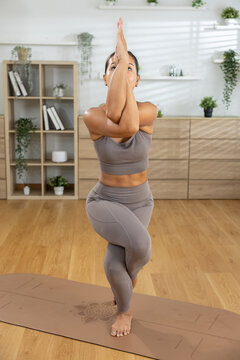 Focused calm young Asian woman practicing advanced yoga eagle pose balancing barefoot on brown mat inside bright home studio wearing gray activewear feeling peaceful