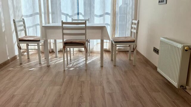 Minimal dining area with white rectangular table, cushioned chairs, light wood floor, sheer curtains, radiator, and natural light creating a clean, modern home setting