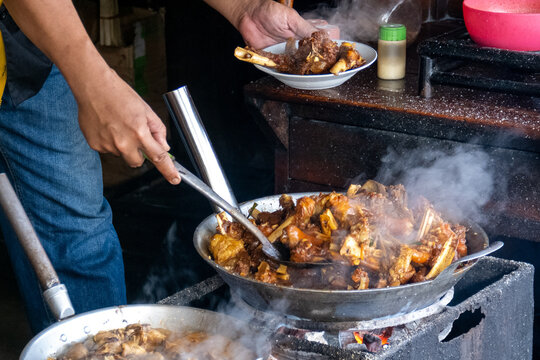 Indonesian street food vendor serving Tengkleng Kambing goat ribs and offal stew into a ceramic plate with a ladle. Hot steam rising from the traditional Javanese food.
