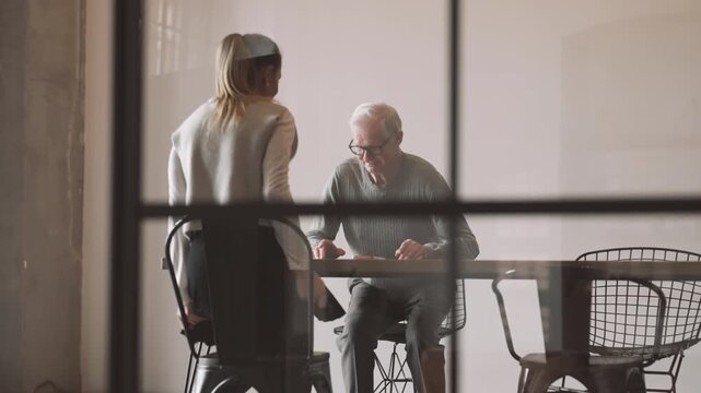 Old man greeting young adult woman in office, job interview in marketing company . Female candidate and male boss shaking hands and sitting down at table in conference hall in modern office, business