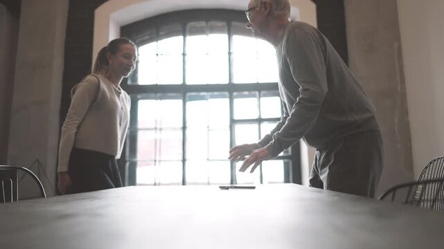 Business meeting for job interview in office, old male boss and female employee. Charming young adult lady and aged man shaking hands and sitting down at table, positive and kindly atmosphere at work