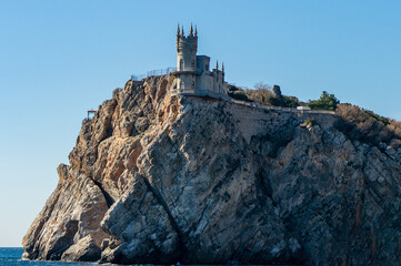 Fototapeta premium Small, intricate Swallow's Nest Castle perched on steep rocky cliff by sea, with clear blue sky in background and winding path leading up rugged terrain. Yalta, Crimea – October 11, 2014