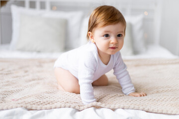 Fototapeta premium a small child girl learns to crawl on a bed in a bright room on cotton white linen, close-up portrait