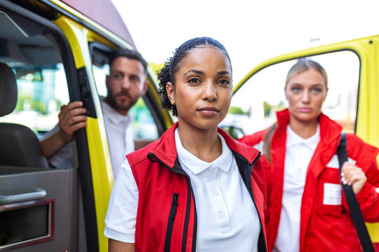 Portrait of female paramedic. Confident emergency medical worker looking at camera with team and ambulance in background, healthcare and medicine concept. Professional first responder at work.