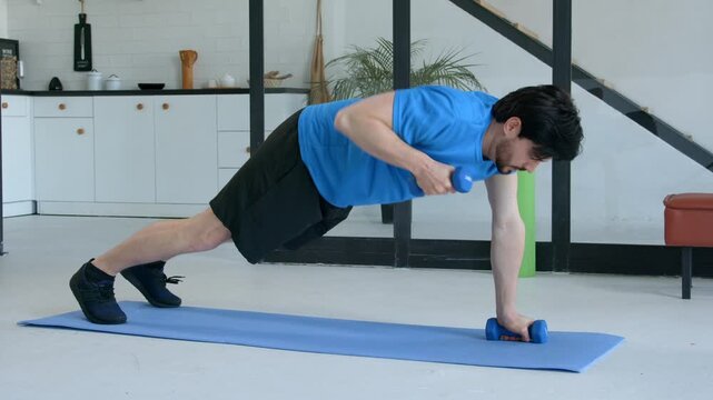 Motivated active sporty fitness man performing dumbbell renegade row exercise, toning obliques, rhomboids, and triceps, developing upper body strength while working out at home.