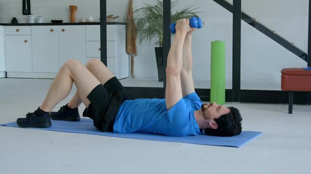 Active fitness attractive man in sportswear lying on yoga mat, practicing dumbbell chest fly exercise, increasing shoulder and core strength while working out at home.