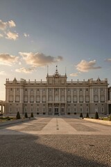 Obraz premium The Royal Palace of Madrid stands majestically in its cobblestone courtyard, surrounded by lush green trees and bathed in warm golden light.