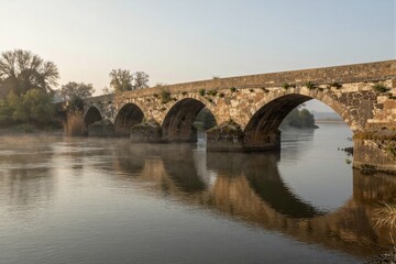 Fototapeta premium A serene stone bridge spans a tranquil river at dawn in a rural landscape surrounded by lush greenery and misty reflections.