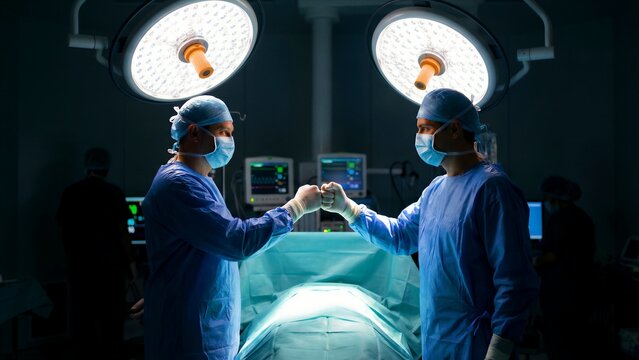 Two dedicated surgeons wearing full surgical attire and sterile masks exchange a supportive fist bump over a brightly lit operating table before a crucial procedure.