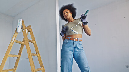 A confident woman of african descent is actively engaged in home improvement tasks. She stands confidently in a room, holding a paintbrush and wearing protective gloves, with a stepladder beside her. © wkproduction