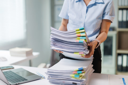 Businesswoman sorting towering stacks of financial reports and administrative files with colorful clips, tackling a chaotic desk of paperwork while managing records and office workflow