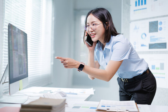 Asian businesswoman talking on a mobile phone while working in a modern office, standing and pointing at content on a desktop computer screen during a busy workday
