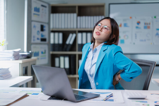 Asian business woman feeling painful backache and discomfort from office syndrome, touching her lower back while sitting at her desk operating a laptop