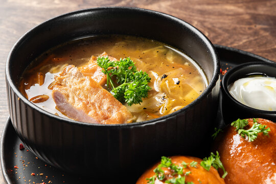 A black bowl of traditional Russian Shchi soup with pork, cabbage, and parsley. Served with garlic pampushki buns and sour cream on a dark wooden background