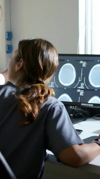 Vertical video: Doctor approaching in coat guiding tech in scrubs at desk, pointing at CT slices