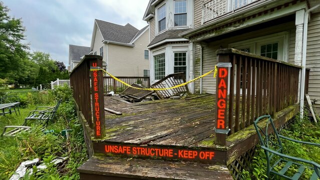 A severely dilapidated and visibly unsafe wooden residential deck, prominently displaying "UNSAFE STRUCTURE - KEEP OFF" and vertical "DANGER" signs, wrapped with bright yellow caution tape, clearly