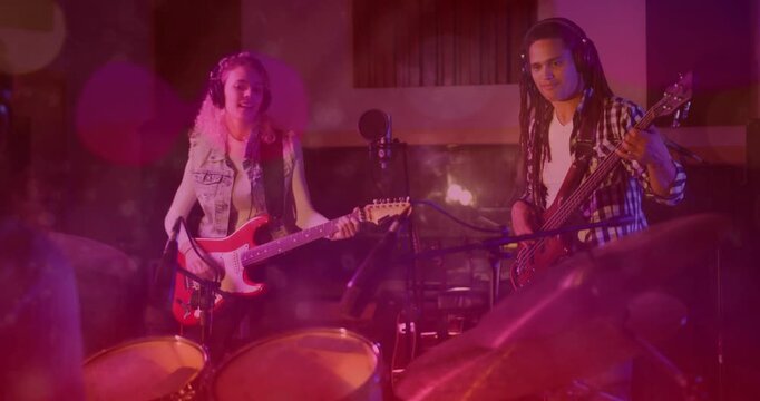 Guitarist initiating strum, bassist responding, rehearsing in studio with drums and purple lighting