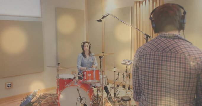 Producer adjusting mic cueing female drummer playing red drum kit in music studio, for recording