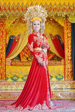 A portrait of a Minangkabau bride in a detailed red and gold traditional dress and elaborate headdress, posed against an ornate wedding backdrop