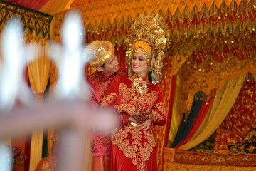 Fototapeta premium A smiling Indonesian couple in traditional red and gold attire on a lushly decorated wedding stage