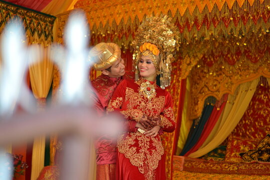 A smiling Indonesian couple in traditional red and gold attire on a lushly decorated wedding stage