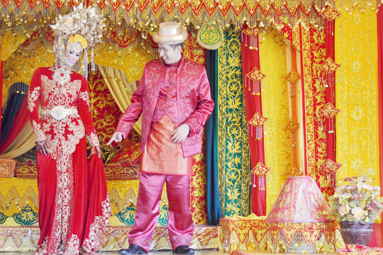A stunning Acehnese couple in traditional red and gold wedding attire, posing elegantly against a vibrant, ornate ceremonial backdrop
