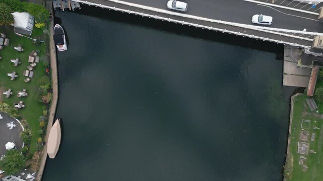 A boat is docked by a grassy area with outdoor seating. A bridge with cars crosses a dark body of water.