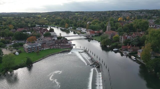 An aerial view shows a wide river with a weir and footbridge, lined by buildings, trees, and a church.