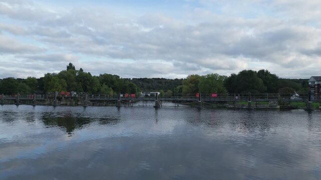 A wide river flows under a cloudy sky, reflecting the surrounding trees and a bridge with "DANGER" signs.
