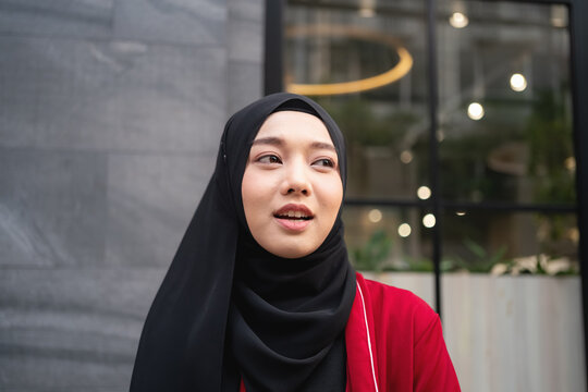 Portrait of a confident young Muslim woman wearing a black hijab and red outfit standing outdoors in a contemporary city setting.