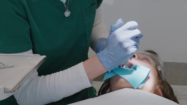 Pediatric orthodontist applies metal clamp with forceps to isolate boy tooth with rubber dam. Preparation stage for endodontic treatment or safe composite restoration in clinic.