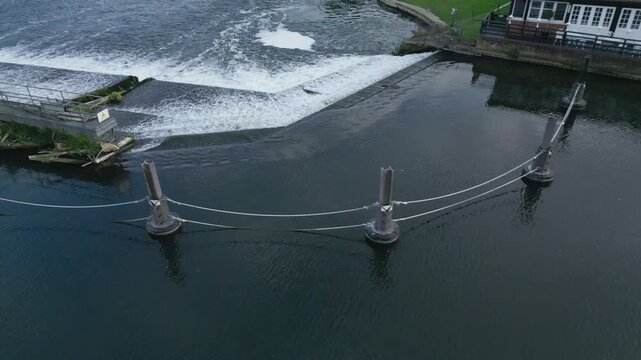 A wide river flows past a building, with a cascading waterfall and a cable barrier in the foreground.