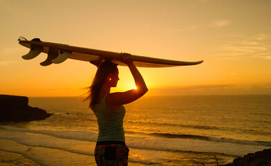 Fototapeta premium SUN FLARE: Young female tourist holding a surfboard on her head at sunset.