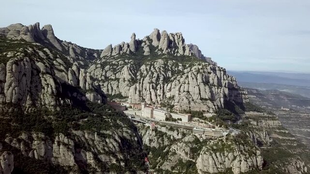 Dramatic aerial pull back reveals the historic monastery nestled within the jagged peaks of the montserrat mountain range in catalonia, spain under a clear sky, showcasing stunn
