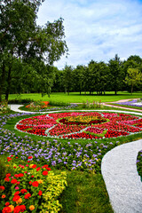 Fototapeta premium Symmetrical circular flower arrangement surrounded by winding paths and greenery, showcasing a harmonious park design under a bright sky.