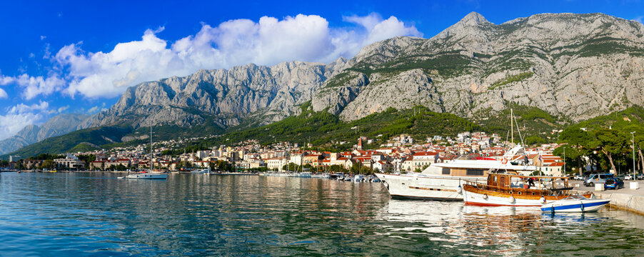 Croatia Makarska Riviera 9K Panorama Scenic Waterfront Promenade with Traditional Boats and Old Town Buildings against Biokovo Mountains Backdrop Adriatic Coast Travel Destination