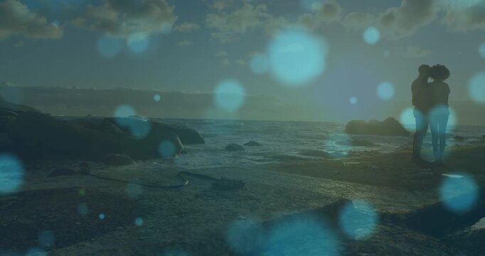 Blue spots of light against african american couple standing together on the rocks near the sea