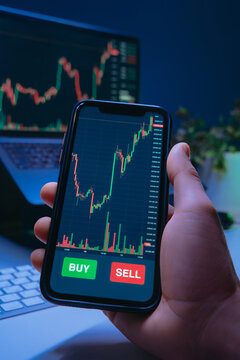 Close-up of a trader hand holds a smartphone with trading app, financial charts or cryptocurrency prices on screen, showing buy and sell buttons during online stock buying or crypto investing.