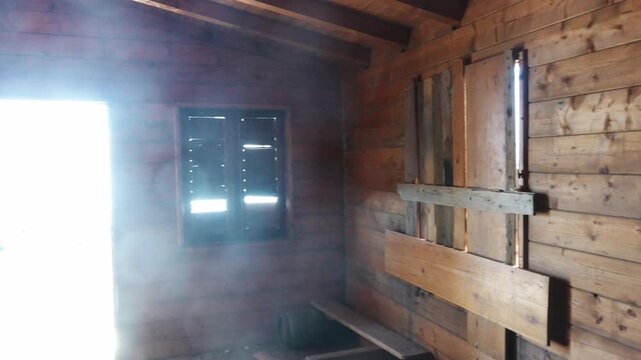 Pan right to left inside a wooden hut toward an open door with sun glare while broken shutters frame dry black lava ground outside.