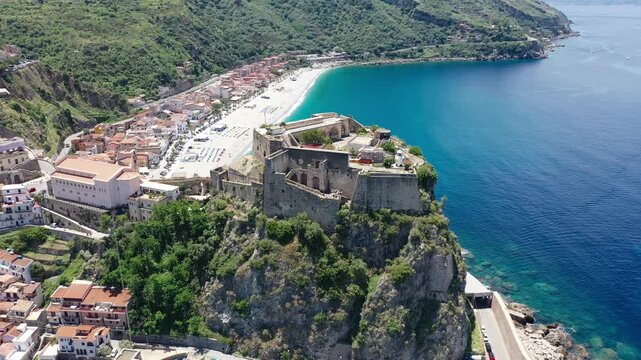 The camera drifts slowly around a castle on a headland in Scilla, with town houses, a church, the coast, and deep blue water behind.